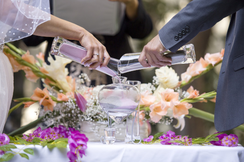 Two well-dressed people pour different colored liquids into the same glass as a commitment ceremony.